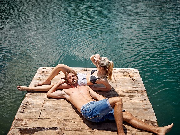Hübner Sonnenbrand und kleine Wunden Mann und Frau liegen auf einem Holzsteg am Wasser und sonnen sich